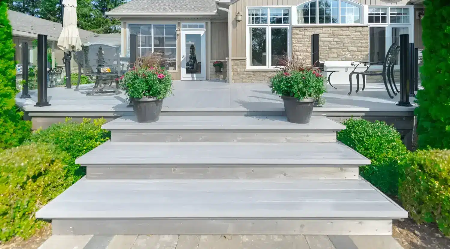 Close-up of Alumadeck light gray aluminum decking featuring wide steps, dark planters, and a contemporary glass railing system on a residential patio.